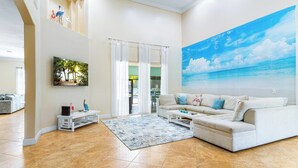 A spacious living room with a large white sofa, a coffee table, and a painting of a beach scene. - Private Villa | Heated Pool + Hot Tub + Sleeps 20 (Port St. Lucie)