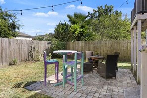 A patio with a table and chairs, a wooden fence, and greenery in the background.