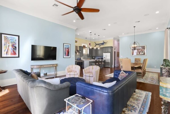 A spacious living room with a large flat-screen TV, a ceiling fan, and a dining area with chairs and a table.