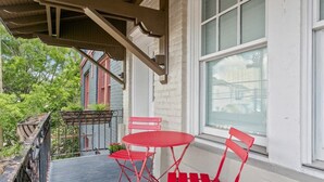 Un porche techado con una mesa y sillas de metal rojo, una ventana y vista de un edificio rojo al fondo.