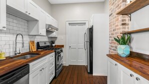 Une cuisine compacte avec des meubles blancs, un mur d’accent en briques, un plan de travail en bois et un réfrigérateur encastré.