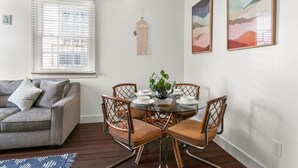 Un salon moderne avec un canapé gris, une table à manger en verre et quatre chaises, ainsi qu’une décoration murale en macramé.