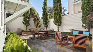 A patio with a table and chairs, surrounded by tall trees and a white wall.