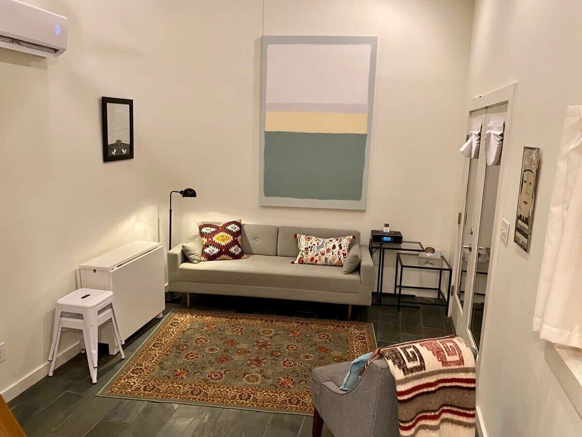 A small, well-lit living room with a sofa, a small table, a radiator, and a patterned rug.