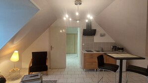 A modern loft apartment with a sloped ceiling, a dining area with a table and chairs, a kitchen with a stove and microwave, and a living area with a sofa and a lamp.