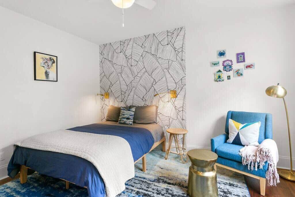 A modern bedroom with a bed, a blue armchair, a small stool, and a wall with a geometric pattern.