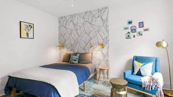 A modern bedroom with a bed, a blue armchair, a small stool, and a wall with a geometric pattern.