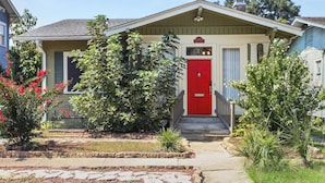 Una casa de un piso con puerta principal roja, un porche pequeño y un jardín delantero bien cuidado con diversas plantas.