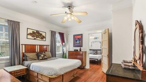 Un dormitorio con una cama de madera, un escritorio, un aparador, un ventilador de techo y una puerta que da a otra habitación.