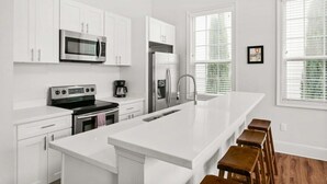 Una cocina moderna con armarios blancos, electrodomésticos de acero inoxidable, una isla grande con taburetes y ventanas amplias con persianas.