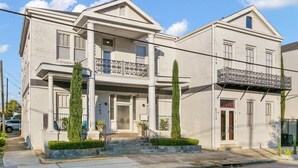Un edificio de dos pisos con un balcón en el segundo nivel, una entrada principal y un pequeño jardín con un árbol alto y esbelto.