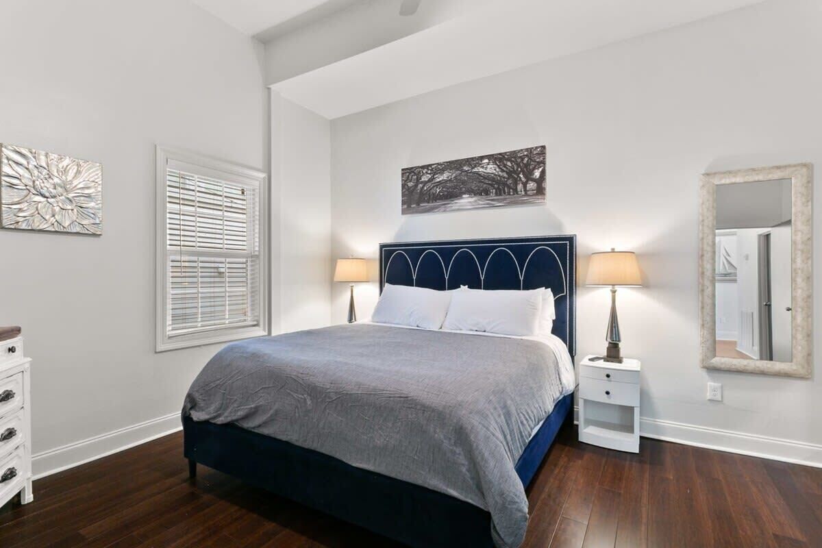 Un dormitorio con cama, lámparas de noche, mesita de noche, cómoda, ventana con persianas y un espejo montado en la pared.