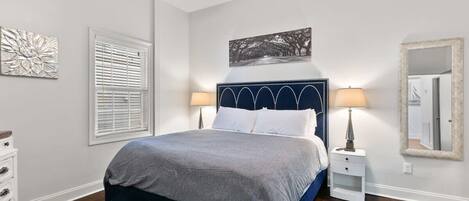 Un dormitorio con cama, lámparas de noche, mesita de noche, cómoda, ventana con persianas y un espejo montado en la pared.