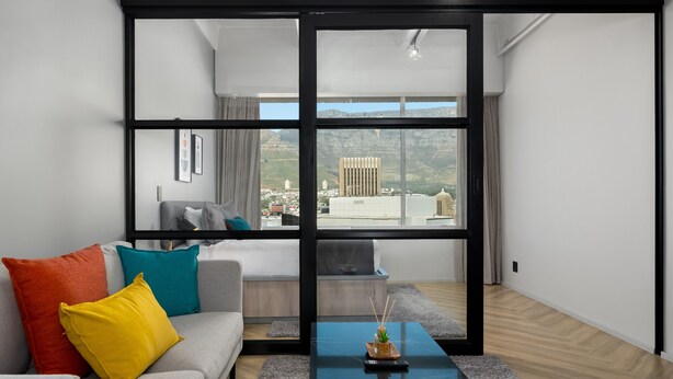 A modern living room with a grey sofa, colorful cushions, a black coffee table, and a large window offering a city view.