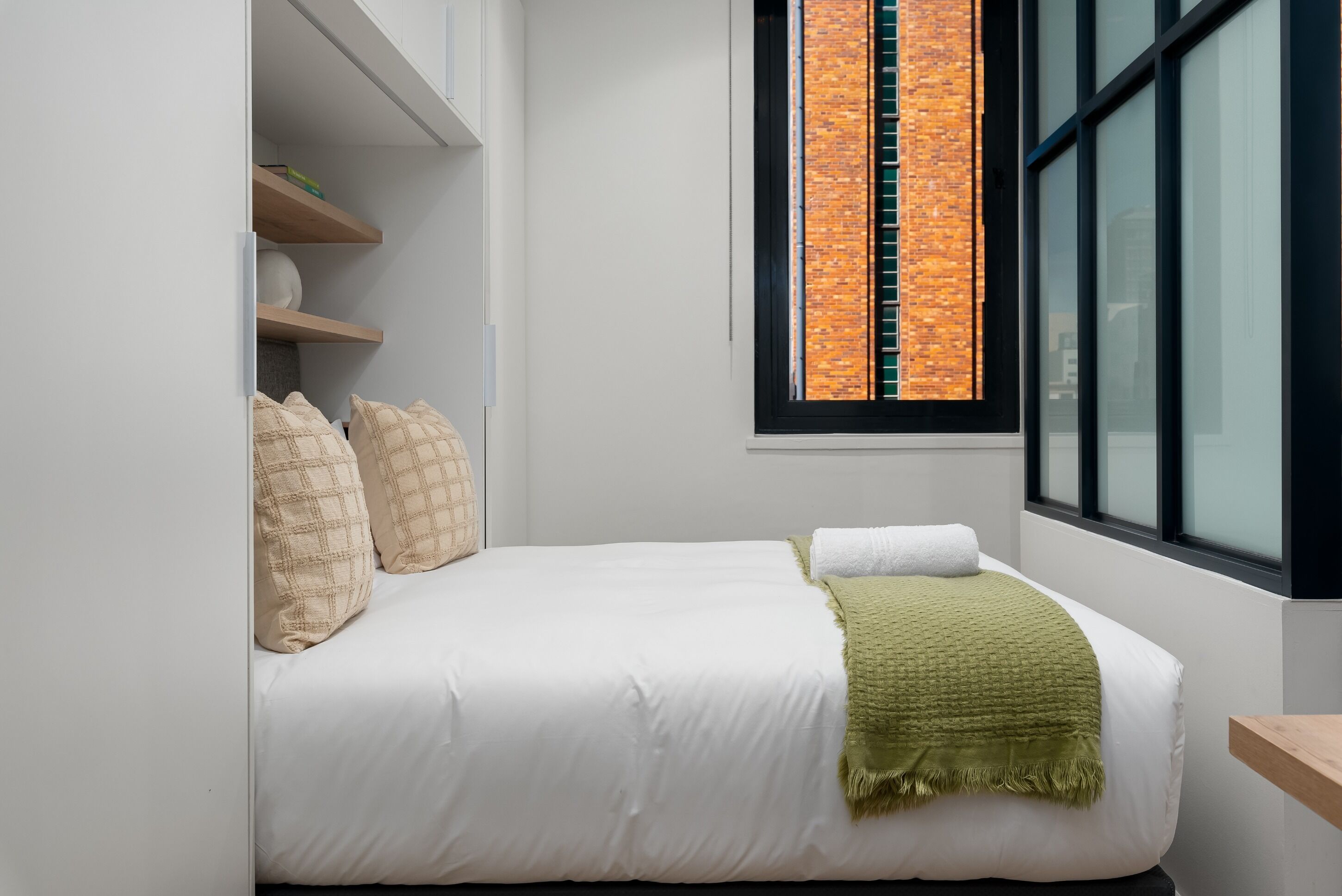 A bedroom with a bed, a window with a brick wall view, and built-in shelves.