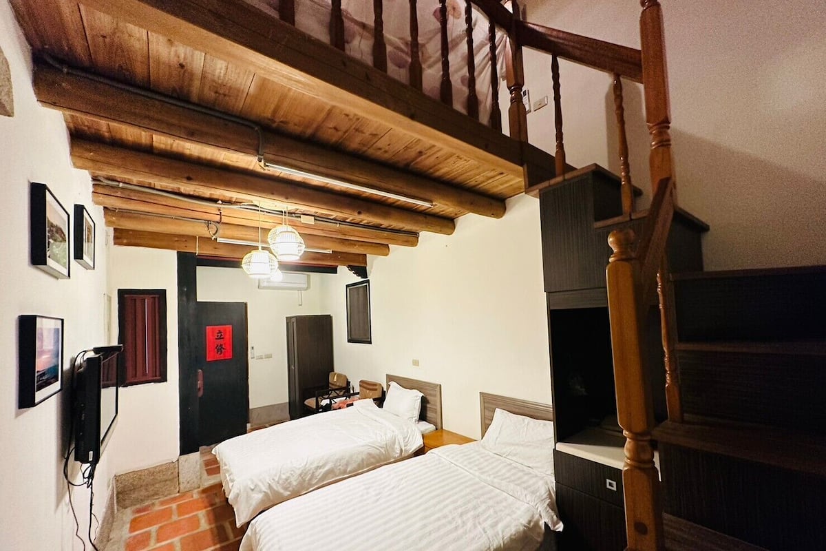 Quarto com duas camas, vigas de madeira no teto, parede de tijolos e uma escada.