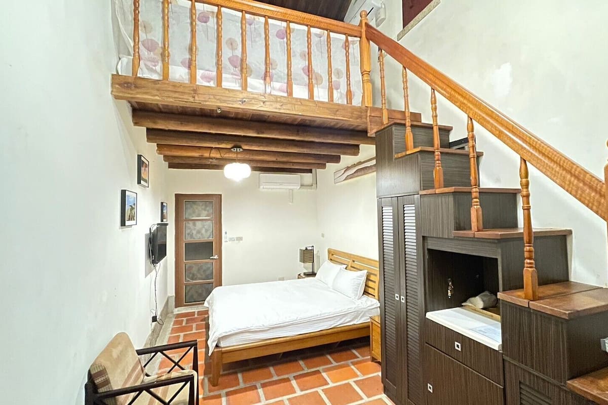 Um quarto com cama de madeira, uma escrivaninha pequena, uma cadeira, um guarda-roupa e uma escada.