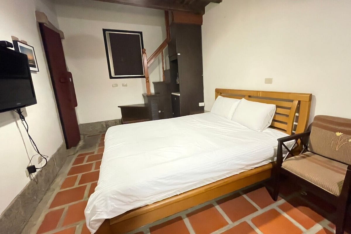 Uma cama de solteiro com cabeceira de madeira, uma cadeira com almofada e uma televisão fixada na parede.
