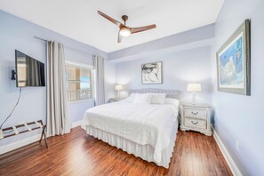 A bedroom with a bed, a ceiling fan, a window with curtains, a bedside table with a lamp, and a wall-mounted TV.