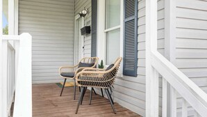 En veranda med två flätade fåtöljer och ett litet bord, ett vitt räcke och ett fönster med fönsterluckor.
