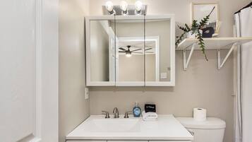 Un baño con lavamanos blanco, espejo, ventilador de techo y retrete.