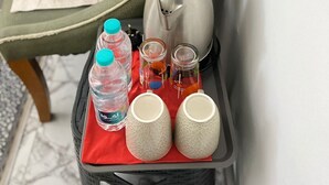 Un carrito pequeño con una tetera, botellas de agua y tazas sobre una bandeja roja, colocado en una silla con cojín.