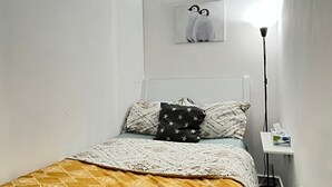 Un dormitorio pequeño y ordenado con cama, lámpara de noche, estante en la pared y un cuadro enmarcado sobre la cama.