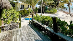 A wooden walkway leading to a yellow building with a thatched roof, surrounded by palm trees and a sandy beach.