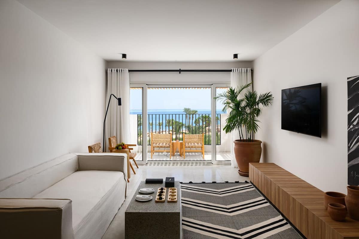 A modern living room with a sofa, a coffee table with decorative items, a large potted plant, a TV mounted on the wall, and a balcony with a view of the ocean.