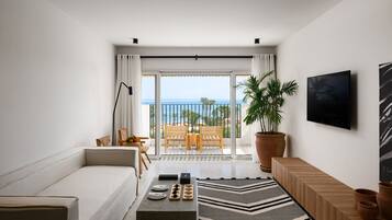 A modern living room with a sofa, a coffee table with decorative items, a large potted plant, a TV mounted on the wall, and a balcony with a view of the ocean.