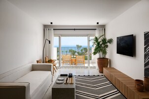 A modern living room with a sofa, a coffee table with decorative items, a large potted plant, a TV mounted on the wall, and a balcony with a view of the ocean.