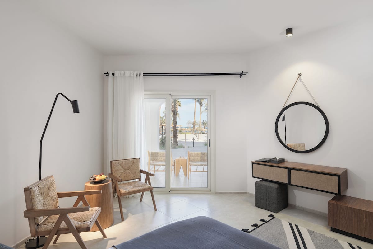 A modern bedroom with a bed, two chairs, a desk, and a large window with a view of palm trees.
