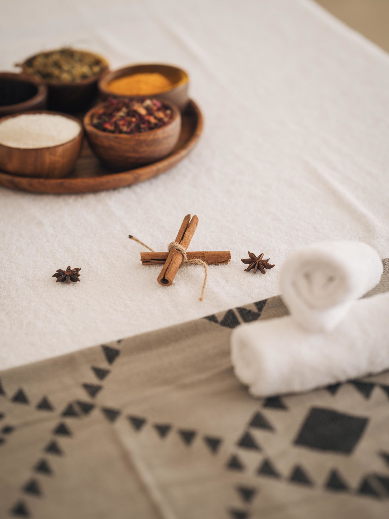 A spa setting with a wooden tray of various bath salts, a rolled white towel, and a cinnamon stick on a textured white surface.
