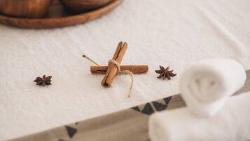 A spa setting with a wooden tray of various bath salts, a rolled white towel, and a cinnamon stick on a textured white surface.