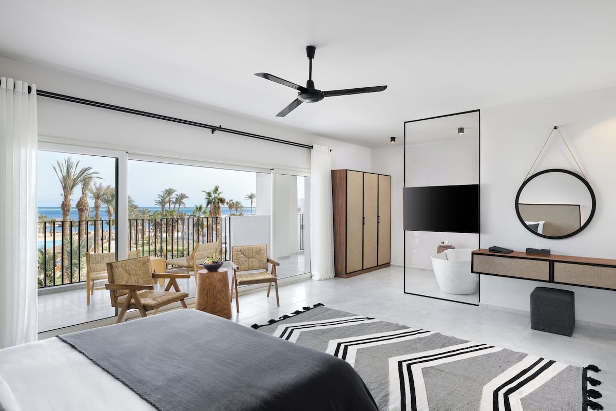 A modern hotel room with a large bed, a ceiling fan, a TV, a wardrobe, a balcony with chairs and a view of the sea.