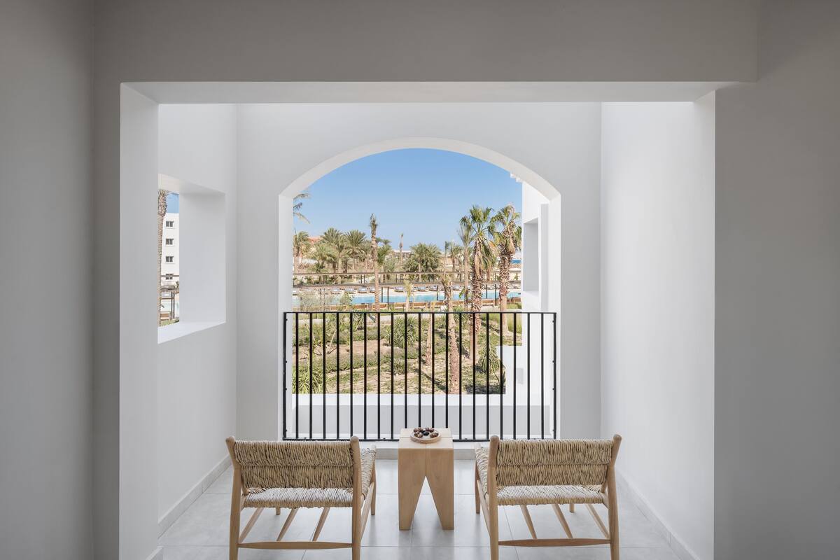 A balcony with two wicker chairs and a small table, overlooking a pool and palm trees.