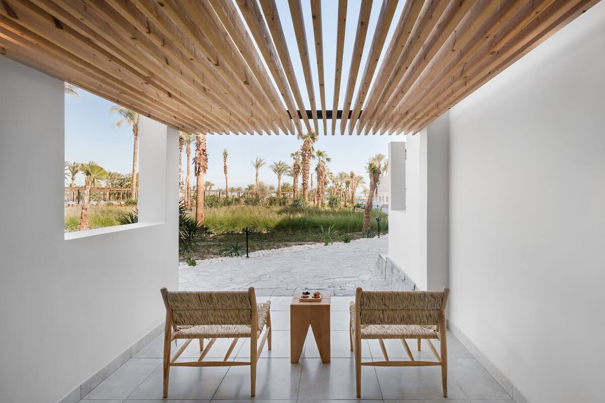 A modern interior with two wicker chairs facing a small wooden table, a wooden ceiling, and a view of a landscaped garden with palm trees.