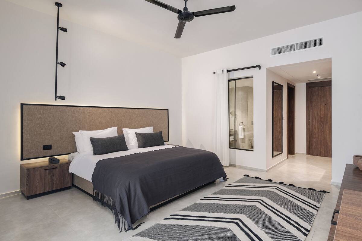 A modern bedroom with a large bed, bedside tables, a ceiling fan, and a view of the hallway.