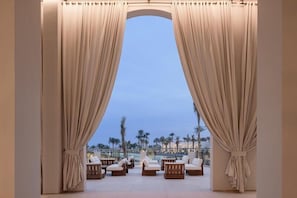 A room with a view of palm trees and a pool, featuring curtains, lounge chairs, and a small table.