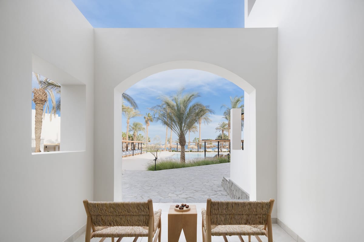 A room with two wicker chairs and a small table, an arched doorway leading to a pool area with palm trees and lounge chairs.