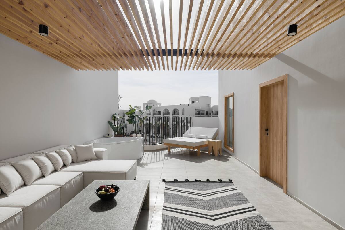A modern outdoor living area with a wooden ceiling, a white sofa, a lounge chair, and a dining table with a bowl of fruit.