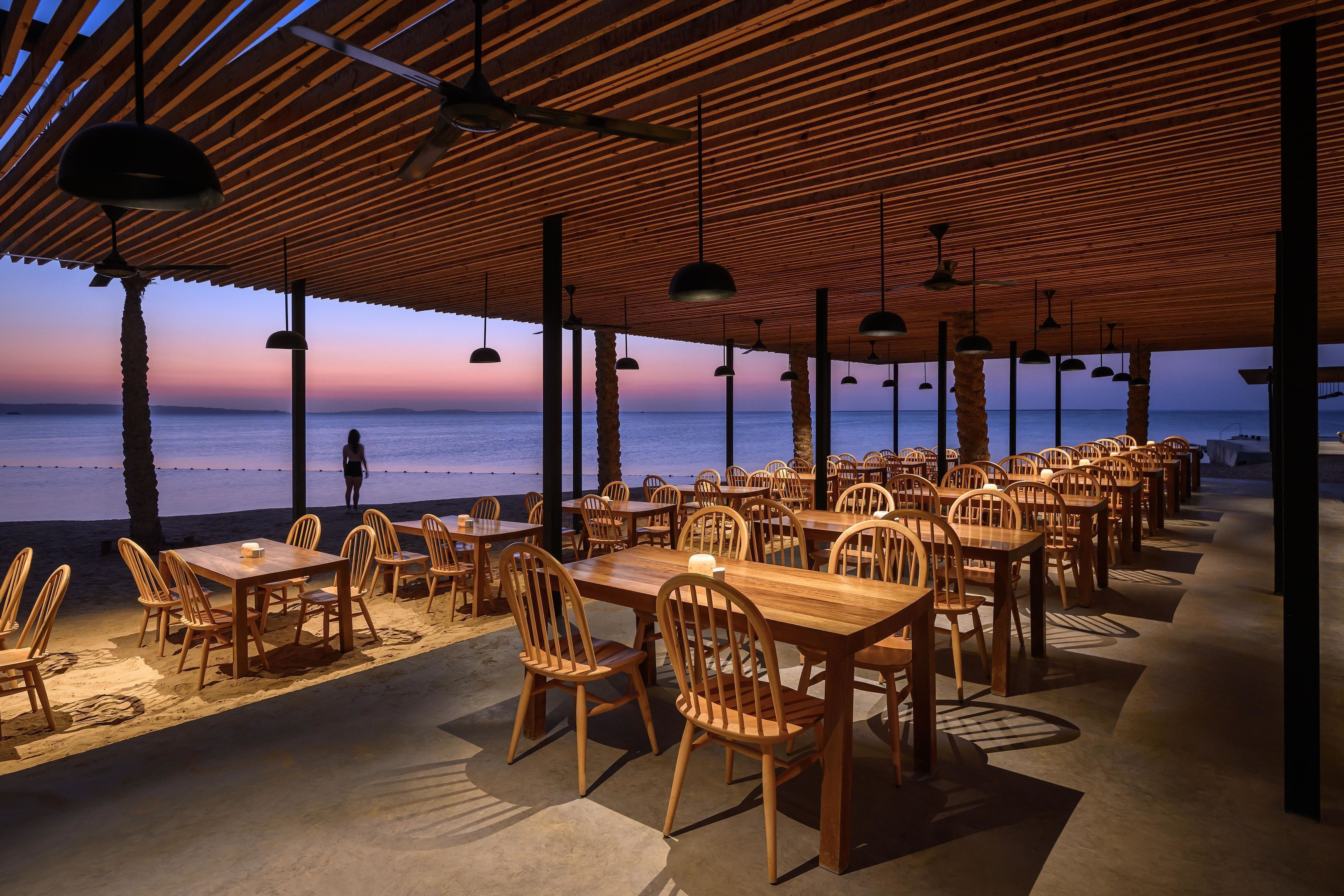 An outdoor dining area with wooden tables and chairs, a covered roof, and a view of the sea.