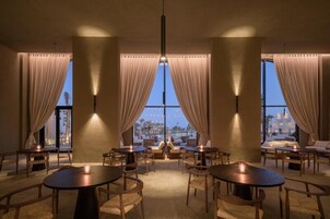 Una habitación con ventanas amplias, cortinas, mesas y sillas dispuestas para comer o conversar.