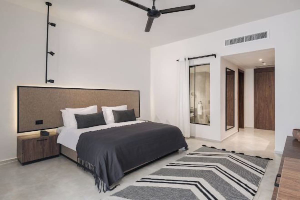 A modern bedroom with a large bed, a ceiling fan, a nightstand, and a view of a bathroom through a glass door.