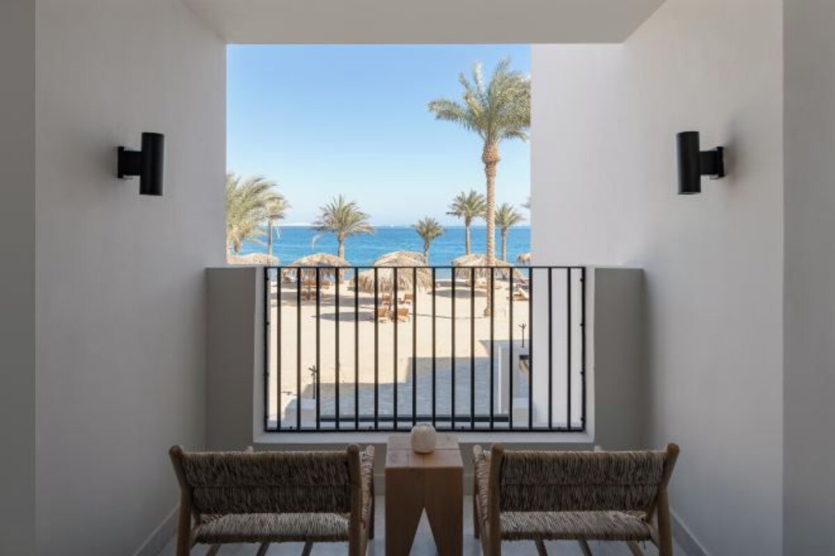 A balcony with two wicker chairs and a small table, overlooking a beach with palm trees and a clear view of the sea.