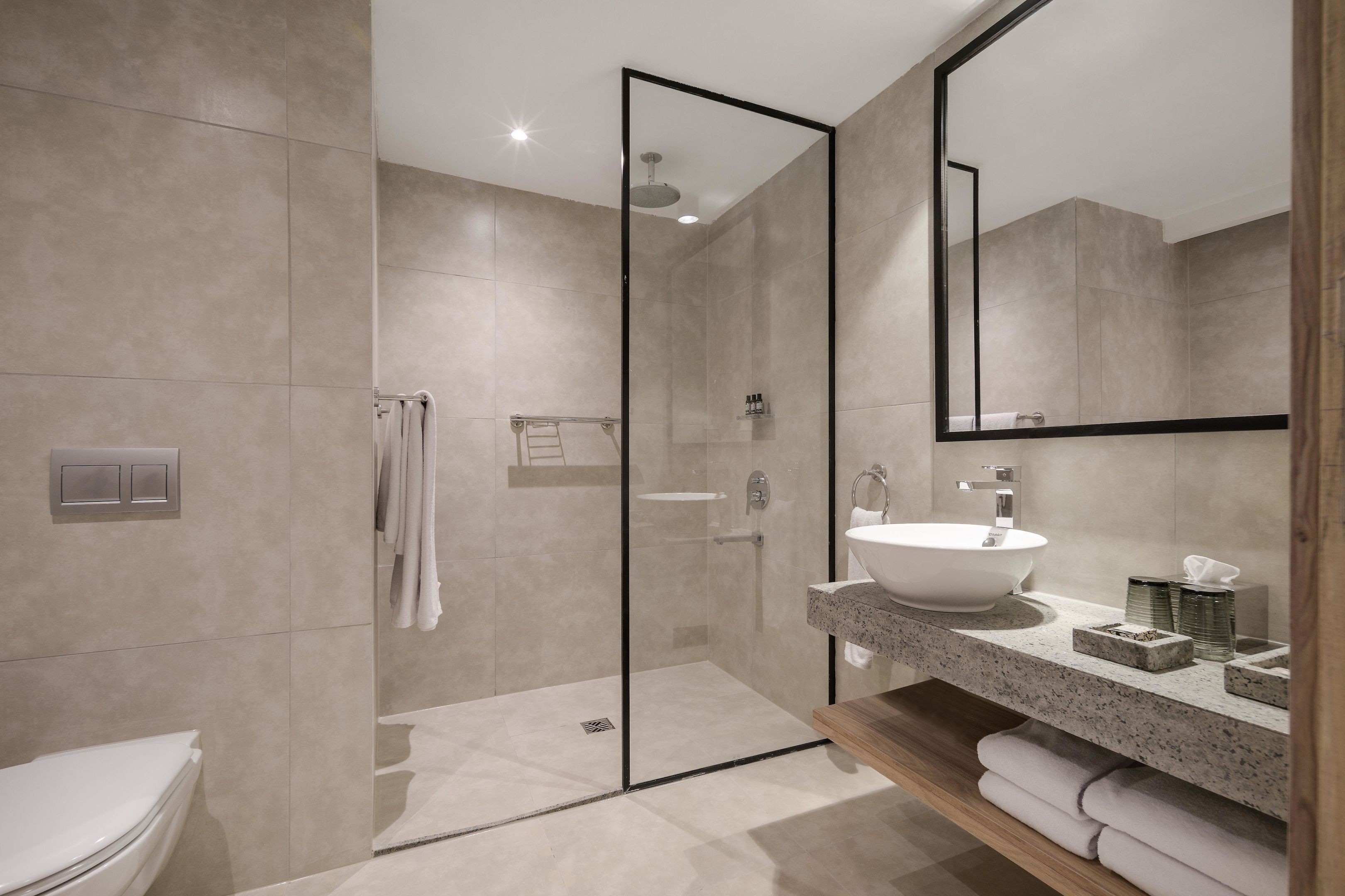 Et moderne badeværelse med en glasvæg omkring brusebadet, et stort spejl og en bordplade i granit med vask og toiletartikler.