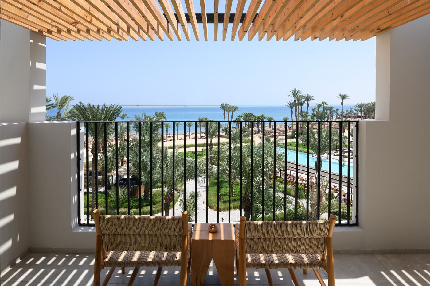A balcony with two wicker chairs and a small table, overlooking a pool and palm trees.