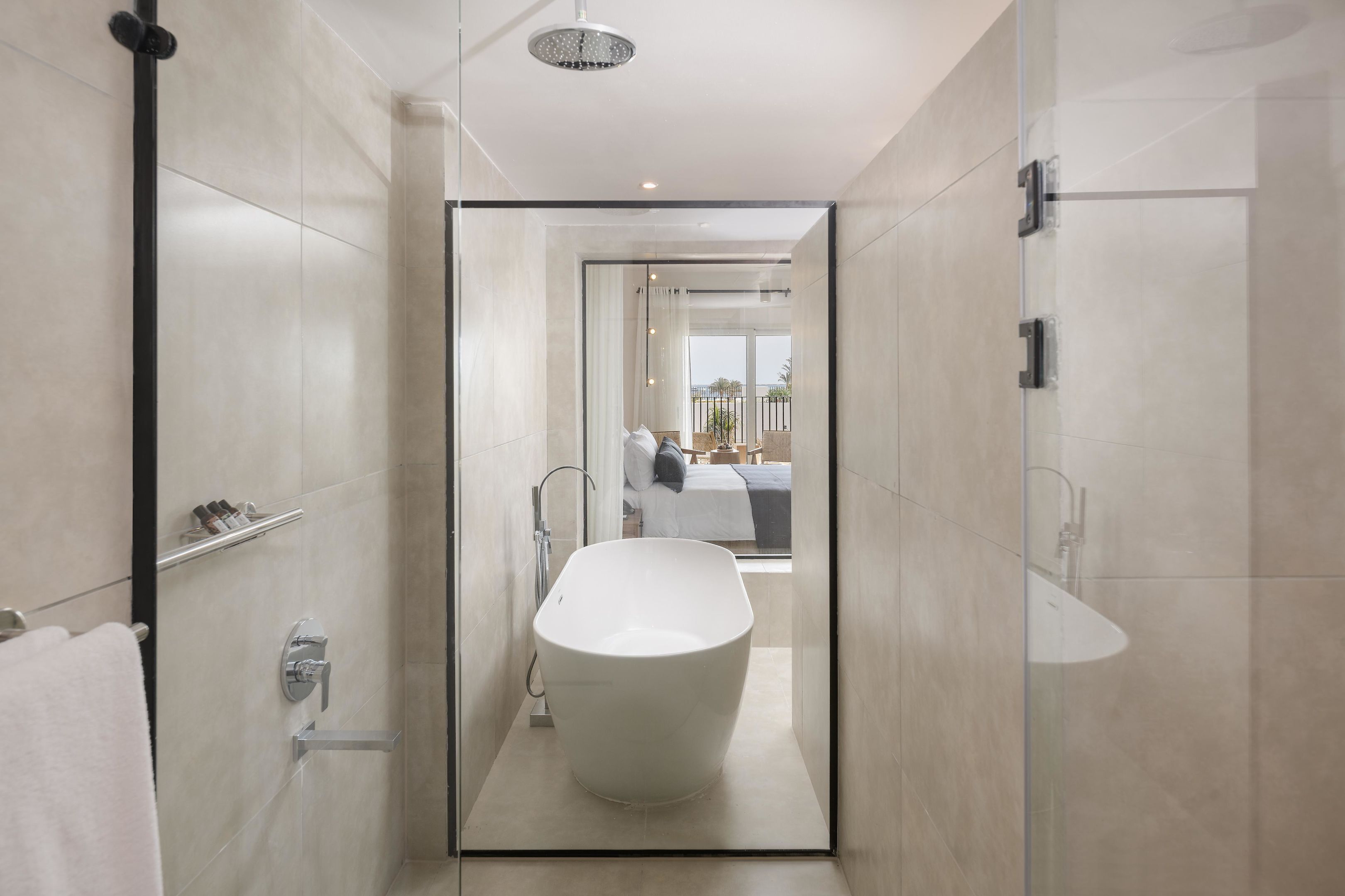 A modern bathroom with a freestanding bathtub, glass shower enclosure, and a visible bedroom with a bed and window.