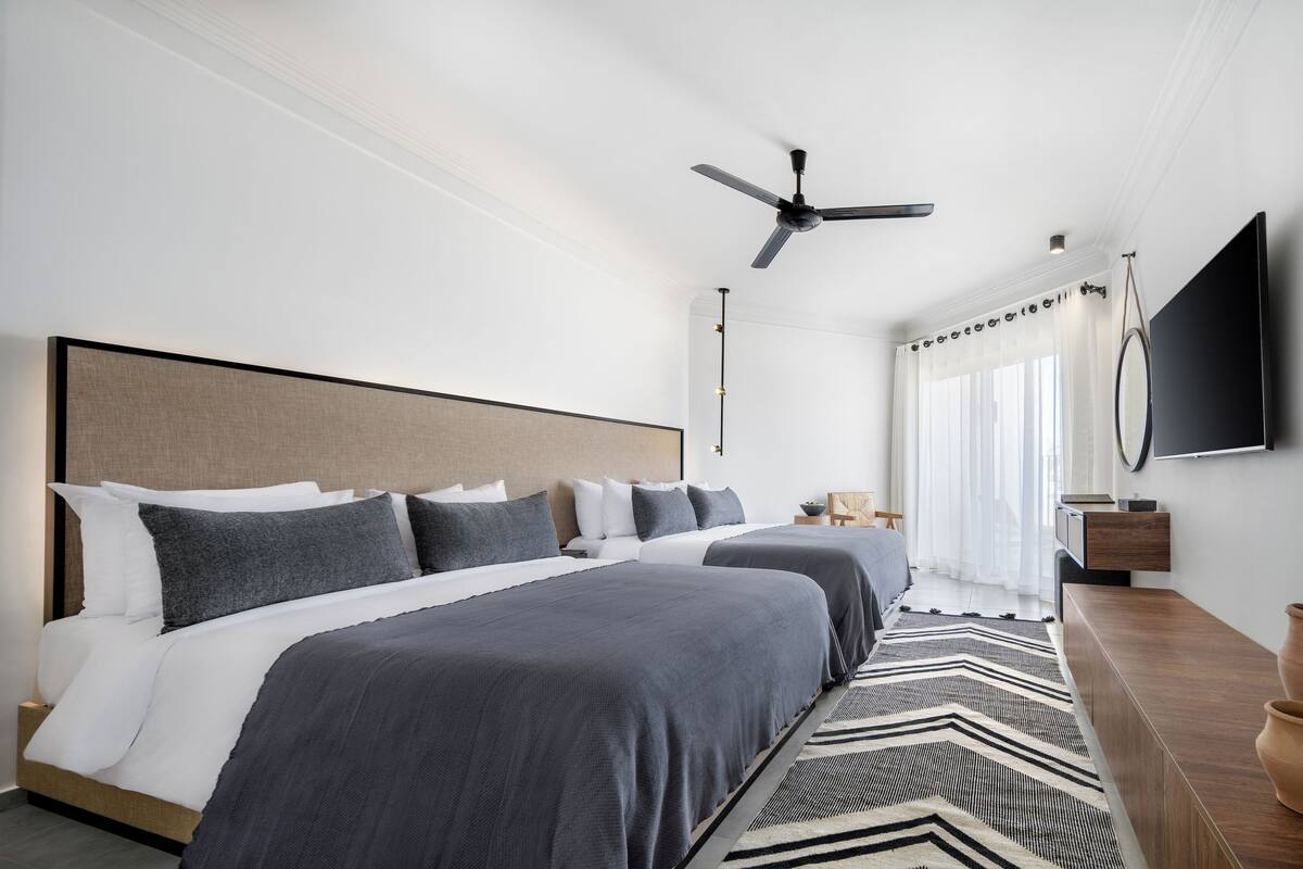 A modern hotel room with a large bed, a ceiling fan, a flat-screen TV, and a large window with sheer curtains.