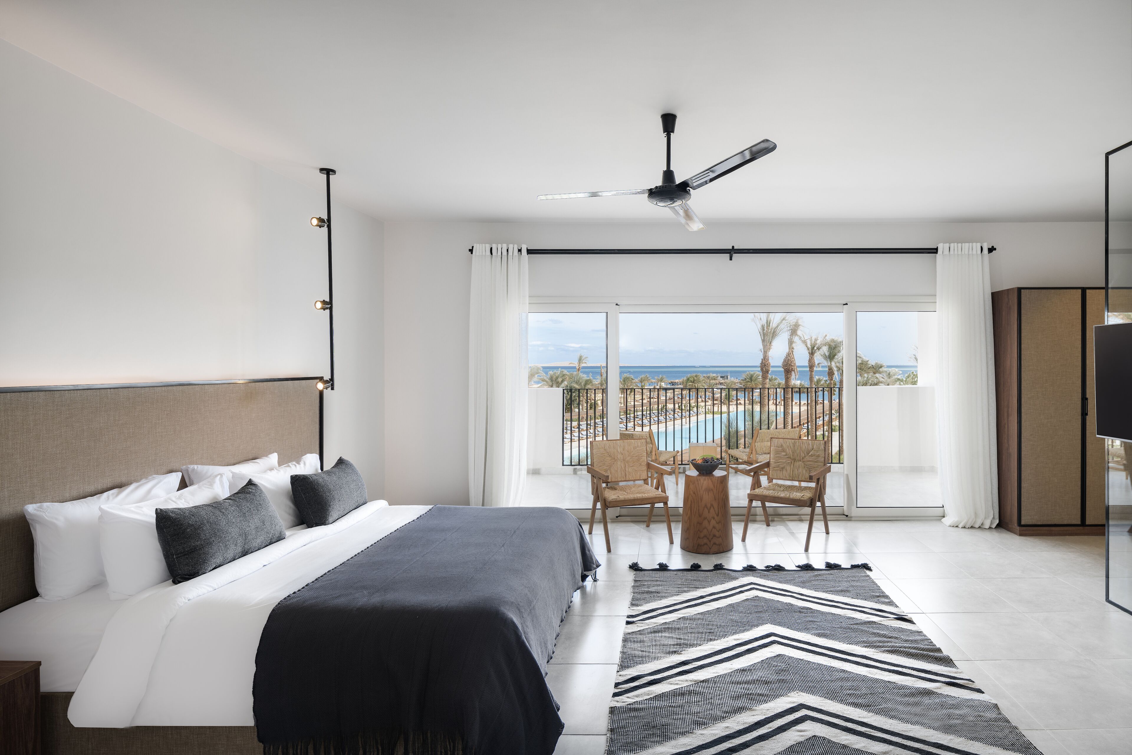 A modern hotel room with a large bed, a ceiling fan, a view of the ocean, and a balcony with chairs and a table.